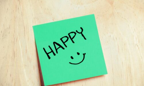 Zielona karteczka samoprzylepna z napisem „HAPPY” i rysunkiem uśmiechniętej buźki na drewnianym stole.
