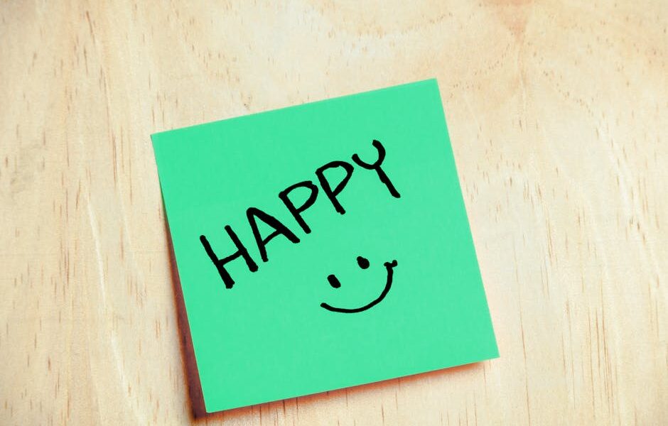 Zielona karteczka samoprzylepna z napisem „HAPPY” i rysunkiem uśmiechniętej buźki na drewnianym stole.