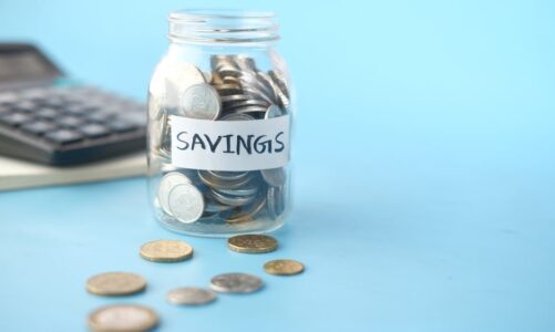 Szklany słoik z napisem „SAVINGS” wypełniony monetami na niebieskim tle obok kalkulatora.