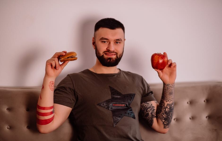 Mężczyzna z tatuażami trzyma w dłoniach hamburgera oraz czerwone jabłko.