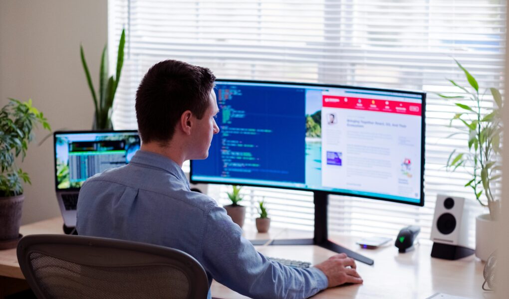 mężczyzna w niebieskiej koszuli siedzący przy biurku przed ultrawide monitorem