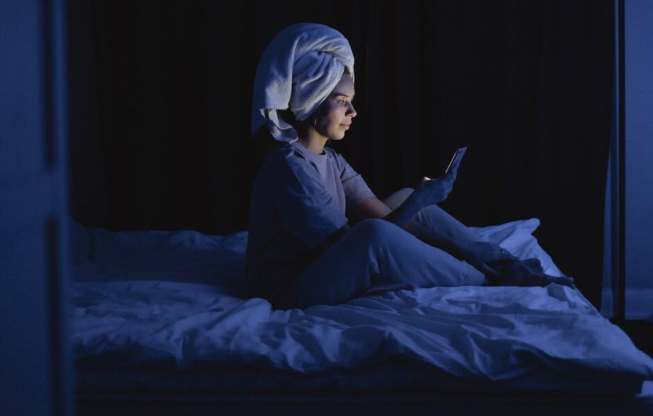 Kobieta w ręczniku na głowie siedzi wieczorem na łóżku i patrzy w telefon.